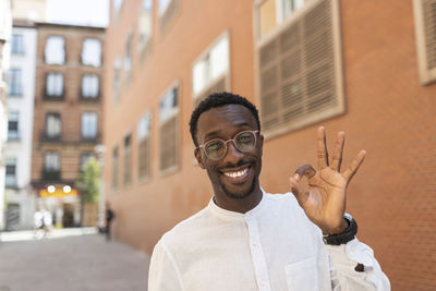 Smiling young man gesturing ok sign in front of buildings