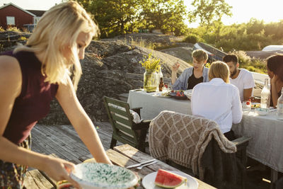 Woman picking watermelon slice while friends are enjoying food on picnic table at harbor