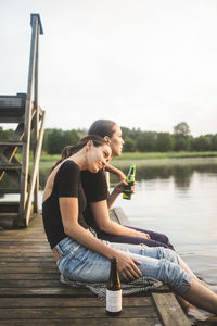 Side view of female friends looking at lake while sitting on jetty during sunset