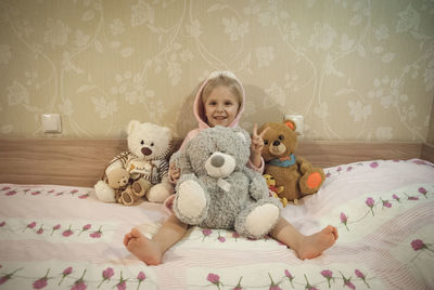 Portrait of cute girl with various teddy bears gesturing peace sign on bed at home