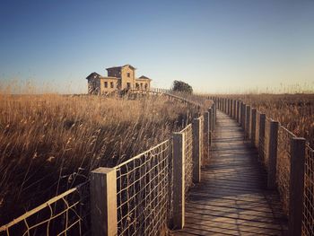 Wooden walkway between fields and blue skies that leads to an abandoned house