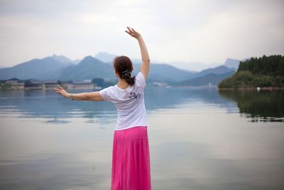 Rear view of young woman dancing while standing at lakeshore against sky
