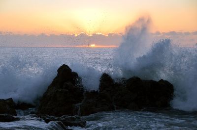 Waves splashing on rocks against sky during sunset