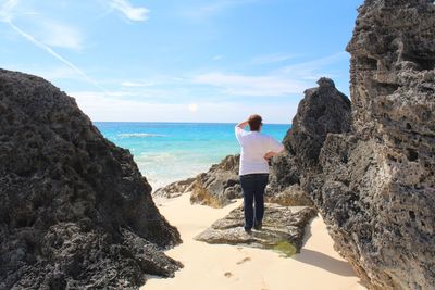 Full length rear view of woman standing on rock at beach against sky