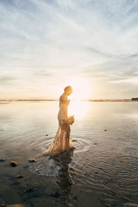 Side view of bride standing at beach against sky during sunset