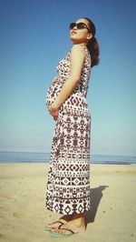 Full length side view of pregnant woman with hands on stomach standing at beach