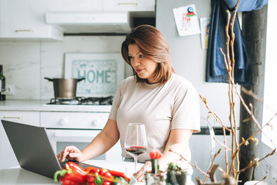 Beautiful smiling brunette young woman plus size body positive using laptop in kitchen at home