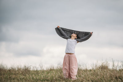 Girl holding sweater while standing on field against cloudy sky at park