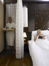Man holding tray by curtain with woman relaxing on bed in hotel room