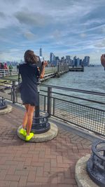 Rear view of woman on retaining wall by sea against cityscape