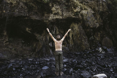 Rear view of shirtless hiker with arms raised standing against mountain