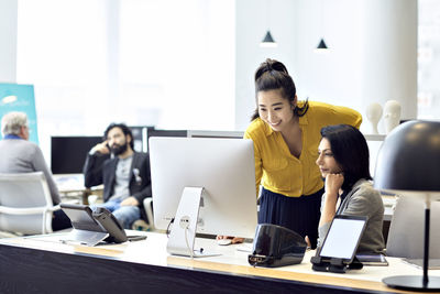 Businesswomen working on desktop computer with male colleagues sitting in back ground