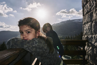 Siblings leaning on railing against cloudy sky