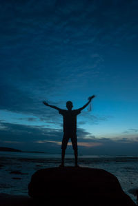 Silhouette man with arms outstretched standing on rock at beach against sky during sunset