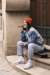 Full body of cheerful female in casual wear and hat listening recording audio message on smartphone while sitting on skateboard near building