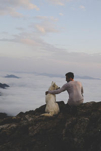 Rear view of man with dog sitting on rock against sky