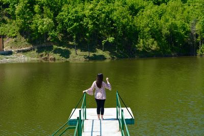 Rear view of young woman standing on pier over lake against trees