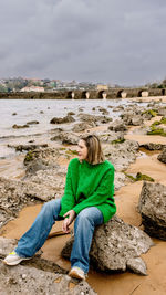 Young woman sitting on rock at beach against sky