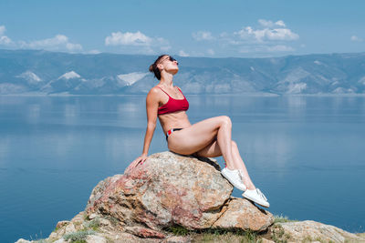 Russian girl in a red swimsuit and sneakers posing sitting on a stone on the background of the lake