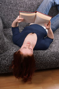 High angle view of young woman reading book while lying down on sofa at home