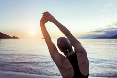 Back view of fit male swimmer in swimsuit and cap standing on seashore and stretching arms before training