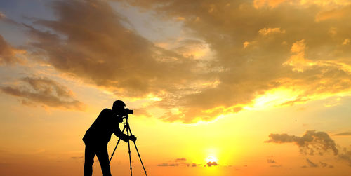Silhouette man photographing on camera against orange sky 
