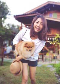 Low angle portrait of smiling woman carrying dog while standing against house