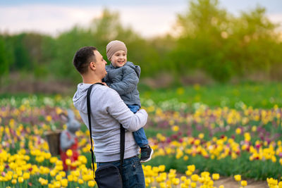 A happy father holds his smiling three-year-old son in hands in a tulip field on a spring day.
