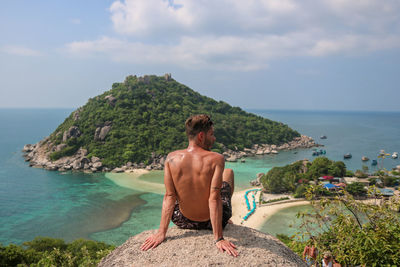 Rear view of shirtless young man looking at sea while sitting against sky