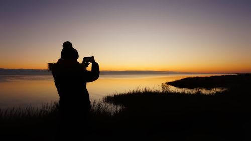 Silhouette woman photographing sea against orange sky