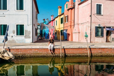 Woman sitting by canal against multi colored buildings