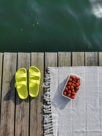 Neon sandals, flip flops with beach rug and box of strawberries on wooden floor beside lake water.