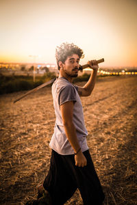 Young man holding sword while walking on field against sky during sunset