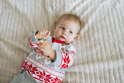 Cute little baby girl in warm overall with garland light in hands lying on the bed