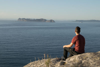 Rear view of man sitting on rock by sea against sky