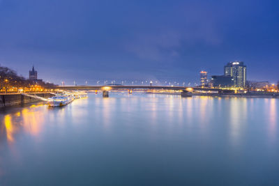 Illuminated bridge over river by buildings against sky at dusk