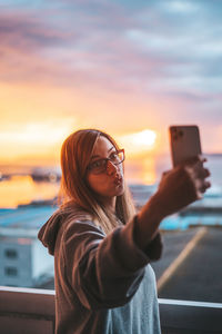 Young woman taking selfie with mobile phone in balcony against sky during sunset