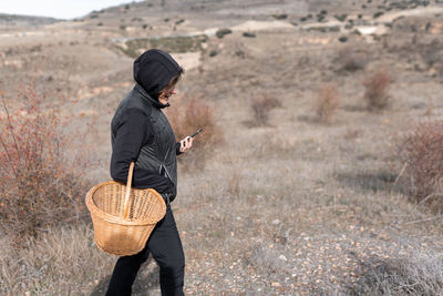 Woman wearing a hoodie is looking at her phone while holding a basket for mushroom picking.