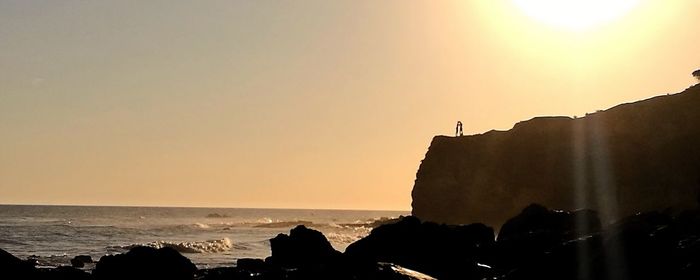 Silhouette cliff by sea against clear sky during sunset