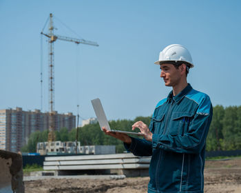 A male builder in a helmet stands against the background of a construction site with a laptop in his