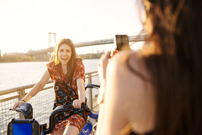 Woman photographing friend on bicycle against clear sky