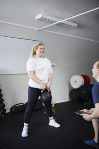 Happy woman exercising with kettlebell in front of fitness instructor at health club