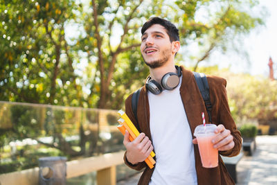 Young man holding drink and books while standing outdoors