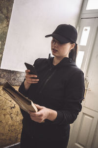 Delivery woman with package using phone while standing against door