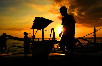Silhouette boats and men at beach against orange sky during sunset