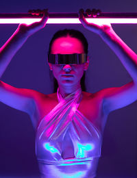 Self assured young female model with long hair in futuristic outfit and vr goggles standing in dark room with neon sword in hands