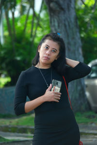 Portrait of young woman using phone while standing against trees