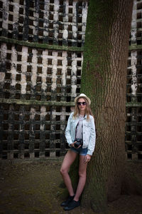 Woman with camera standing by tree trunk against wall