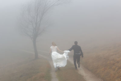 Rear view of newlywed couple running on field during foggy weather
