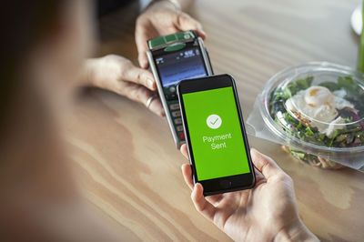 Cropped hands of female customer making mobile payment to owner on wooden table in cafeteria